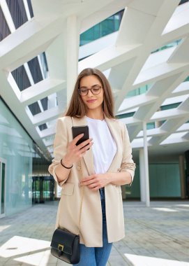 Şehirde yürürken akıllı telefonuna gülümseyen bir kadın. Esmer kadın iş kıyafeti giyiyor..