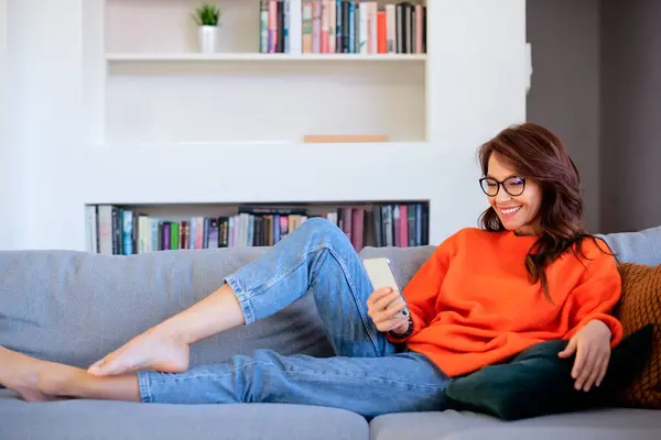 stock image Full length of an attractive brunette haired woman relaxing on the sofa at home. Brunette haired female using smartphone. 