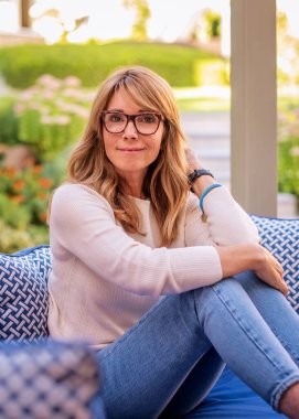 Gülümseyen orta yaşlı kadın terasta koltukta dinleniyor. Beyaz gömlek ve kot giyen sarışın kadın.. 