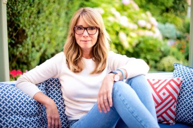 Gülümseyen orta yaşlı kadın terasta koltukta dinleniyor. Beyaz gömlek ve kot giyen sarışın kadın.. 