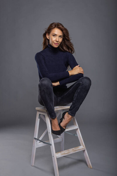 Attractive brown-haired woman sitting in front of a grey wall. Mid aged female wearing black turtleneck sweater and smiling. Studio shot. Isolated background. Full length shot.