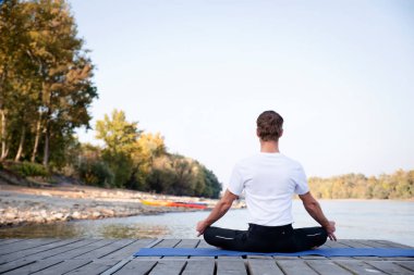 Göl kenarındaki iskelede yoga yapan beyaz bir adam. Meditasyon yapan ve rahatlatan bir adamın arka görünüşü. 