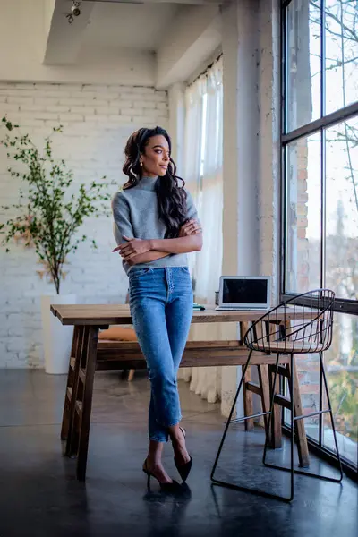 Gülümseyen Afro-Amerikan kadın pencerenin yanında duruyor. Siyah saçlı kadın kazak ve kot pantolon giyiyor. Tam boy çekim. 