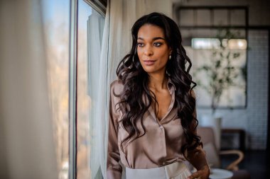 Pencerenin önünde dikilmiş dışarı bakan Afrikalı Amerikalı genç bir kadının yakın plan portresi. Uzun saçlı, günlük kıyafetler giyen bir kadın..