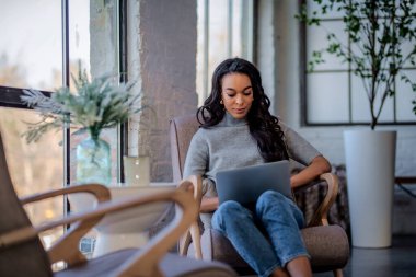 Pencere kenarındaki koltukta oturan ve bilgisayarını kullanan çekici bir Afrikalı Amerikalı kadın. Kafede çalışan gülümseyen iş kadını. Tam boy çekim. 