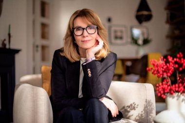 Evdeki koltukta oturan gülümseyen sarı saçlı kadın. Orta yaşlı kadın siyah ceket ve pantolon giyiyor ve kameraya gülümsüyor..