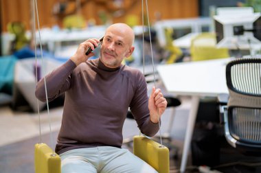 Senior businessman sitting at the office and using his smartphone. Professional male talking with somebody on his phone and wearing business casual.  clipart