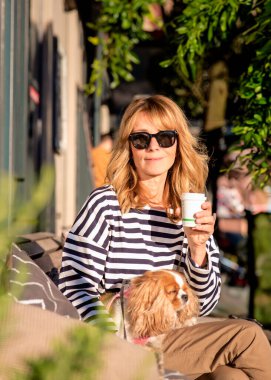 Sarışın, orta yaşlı bir kadın bir kafenin terasında köpeğiyle oturuyor. Gündelik kıyafetler ve güneş gözlüğü takan çekici bir kadın.. 