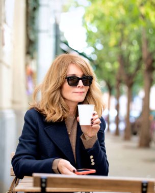 Orta yaşlı bir kadın kafenin dışında bir masada oturuyor ve bir fincan kahve içerken akıllı telefonunu kullanıyor. Gündelik kıyafetler ve güneş gözlüğü takan kendinden emin bir kadın.. 