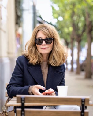 Orta yaşlı bir kadın kafenin dışında bir masada oturuyor ve bir fincan kahve içerken akıllı telefonunu kullanıyor. Gündelik kıyafetler ve güneş gözlüğü takan kendinden emin bir kadın.. 