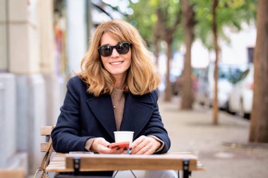 Orta yaşlı bir kadın kafenin dışında bir masada oturuyor ve bir fincan kahve içerken akıllı telefonunu kullanıyor. Gündelik kıyafetler ve güneş gözlüğü takan kendinden emin bir kadın.. 