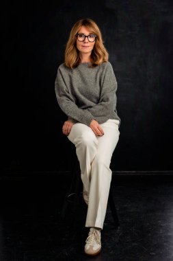 Siyah arka planda çekici bir kadının tam uzunlukta stüdyo portresi. Sarı saçlı, gri kazaklı ve beyaz pantolonlu bir kadın. Kameraya bakıyor ve gülümsüyor. Boşluğu kopyala.