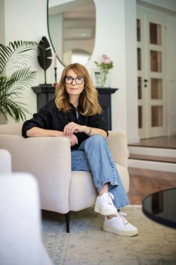 Orta yaşlı, çekici bir kadın evdeki koltukta dinleniyor. Sarı saçlı, gözlüklü ve neşeli gülümseyen bir kadın. Tam boy çekim. 