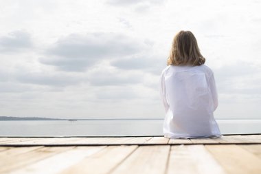 Göl kenarındaki iskelede oturan sarışın, orta yaşlı bir kadının arka görüntüsü. Tam yol ileri. Beyaz gömlek ve kot pantolon giyen kendinden emin bir kadın..