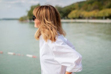 Gölün kenarında duran sarı saçlı bir kadının portresi. Güneş gözlüğü ve beyaz gömlek giyen orta yaşlı bir kadın. Yakın çekim..