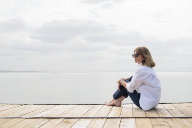 Göl kenarındaki iskelede oturan sarışın, orta yaşlı bir kadın. Tam yol ileri. Beyaz gömlek ve kot pantolon giyen kendinden emin bir kadın..