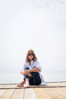 Göl kenarındaki iskelede oturan sarışın, orta yaşlı bir kadın. Tam yol ileri. Beyaz gömlek ve kot pantolon giyen kendinden emin bir kadın..