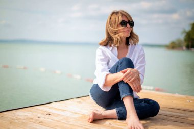 Göl kenarındaki iskelede oturan sarışın, orta yaşlı bir kadın. Tam yol ileri. Beyaz gömlek ve kot pantolon giyen kendinden emin bir kadın..