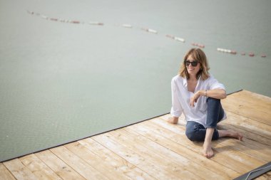 Göl kenarındaki iskelede oturan sarışın, orta yaşlı bir kadın. Tam yol ileri. Beyaz gömlek ve kot pantolon giyen kendinden emin bir kadın. Yüksek açılı çekim. 