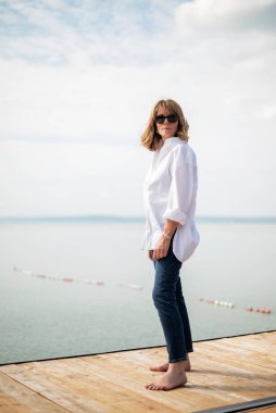 Göl kenarındaki iskelede duran sarışın, orta yaşlı bir kadın. Tam yol ileri. Beyaz gömlek ve kot pantolon giyen kendinden emin bir kadın.. 