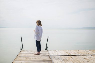 Göl kenarındaki iskelede duran sarışın, orta yaşlı bir kadının arka görüntüsü. Tam yol ileri. Beyaz gömlek ve kot pantolon giyen kendinden emin bir kadın..