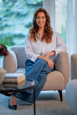 Kumral saçlı çekici bir kadın penceredeki koltukta oturuyor ve kameraya gülümsüyor. Beyaz gömlek ve kot pantolon giyen kendinden emin bir kadın. Tam boy çekim.  