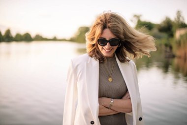 Göl kenarındaki iskelede duran sarışın, orta yaşlı bir kadın. Beyaz takım elbiseli ve güneş gözlüklü çekici bir kadın..  