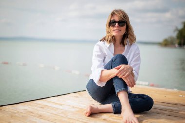 Göl kenarındaki iskelede oturan sarışın, orta yaşlı bir kadın. Tam yol ileri. Beyaz gömlek ve kot pantolon giyen kendinden emin bir kadın.. 