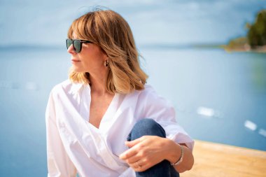 Göl kenarındaki iskelede oturan sarışın, orta yaşlı bir kadın. Beyaz gömlek ve güneş gözlüğü takan kendine güvenen bir kadının yakın çekimi.. 