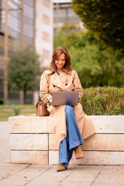 Esmer kadın şehir parkında oturuyor ve iş için dizüstü bilgisayar kullanıyor. Sıradan kıyafetler giyen orta yaşlı mutlu bir kadın. Tam boy çekim.  