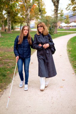 Şehir parkında kadın arkadaşıyla yürüyen görme engelli bir kadın. Görme engelli arkadaşına yardım eden olgun, beyaz bir kadın. Tam uzunluk.  