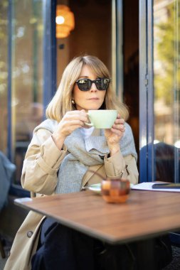 Orta yaşlı bir kadın kafenin dışında bir masada oturuyor ve bir fincan kahve içiyor. Gündelik kıyafetler ve güneş gözlüğü takan kendinden emin bir kadın..  