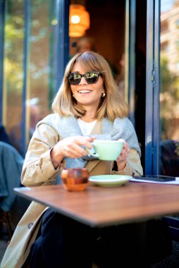 Orta yaşlı bir kadın kafenin dışında bir masada oturuyor ve bir fincan kahve içiyor. Gündelik kıyafetler ve güneş gözlüğü takan kendinden emin bir kadın..  
