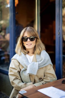 Kafenin dışındaki masada oturan orta yaşlı bir kadın. Sarı saçlı, günlük kıyafetler ve güneş gözlüğü takan bir kadın..  