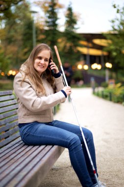 Beyaz baston tutan ve şehirde bir bankta oturan görme engelli bir kadın. Günlük giysiler içinde gülümseyen bir kadın arıyor. Tam boy çekim.  