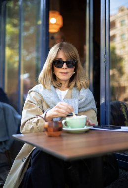 Orta yaşlı bir kadın kafenin dışında bir masada oturuyor ve bir fincan kahve içiyor. Gündelik kıyafetler ve güneş gözlüğü takan kendinden emin bir kadın..  
