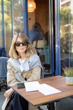 Kafenin dışındaki masada oturan orta yaşlı bir kadın. Gündelik kıyafetler ve güneş gözlüğü takan kendinden emin bir kadın..  