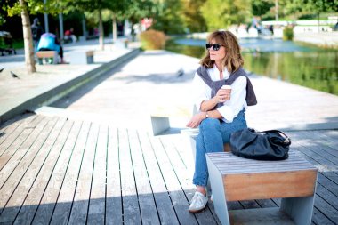 Şehir parkında bankta oturmuş kahvesini içen orta yaşlı bir kadın. Sarışın, beyaz gömlek ve kot giyen ve birini bekleyen bir kadın. Tam boy çekim. 