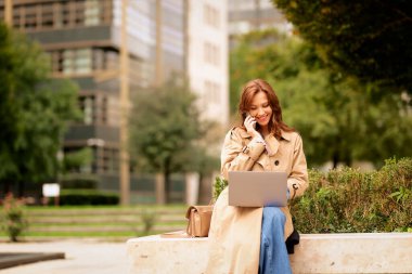 Esmer kadın şehir parkında oturuyor ve iş için dizüstü bilgisayar ve akıllı telefon kullanıyor. Mutlu orta yaşlı kadın günlük kıyafetler giyiyor ve telefonla konuşuyor. Tam boy çekim.  