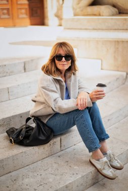 Şehir merkezindeki merdivenlerde oturan orta yaşlı bir kadın kahve içiyor. Tam yol ileri. Sarı saçlı kadın ceket ve kot pantolon giyiyor..  