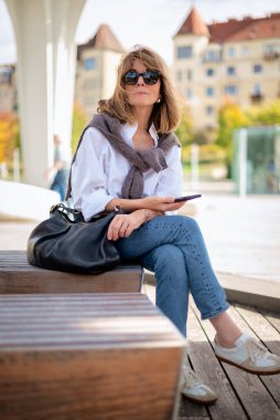 Şehir parkında bankta oturan orta yaşlı bir kadın. Sarışın, beyaz gömlek ve kot giyen ve birini bekleyen bir kadın. Tam boy çekim. 