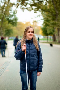 Beyaz bastonuyla parkta yürüyen görme engelli bir kadın. Sarı saçlı, sıradan kıyafetler giyen bir kadın.. 