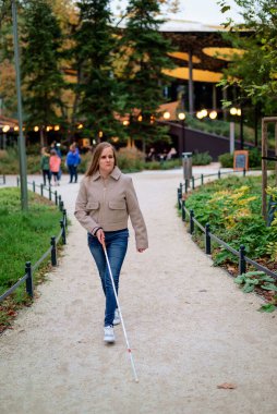 Beyaz bastonuyla parkta yürüyen görme engelli bir kadın. Sarı saçlı, sıradan kıyafetler giyen bir kadın. Tam boy çekim.