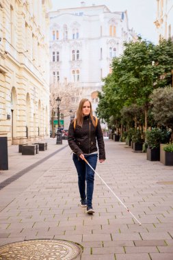 Sokakta yürüyen beyaz bastonlu kör kadın portresi. Sıradan kıyafetler giyen ve bastonuyla sokakta yürüyen görme engelli bir kadın. Tam boy çekim.