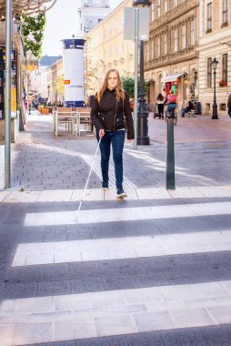 Şehirde yoldan karşıya geçen beyaz bastonlu kör bir kadının portresi. Sıradan kıyafetler giyen ve sopasıyla karşıya geçen görme engelli bir kadın. Tam boy çekim. 