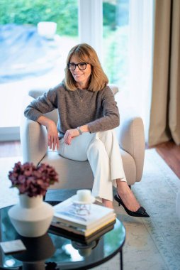 Çekici orta yaşlı kadın evdeki koltukta dinleniyor. Süveter ve pantolon giyen sarışın kadın. Tam boy çekim.  