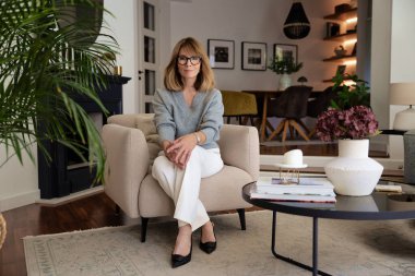 Çekici orta yaşlı kadın evdeki koltukta dinleniyor. Süveter ve pantolon giyen sarışın kadın. Tam boy çekim.  