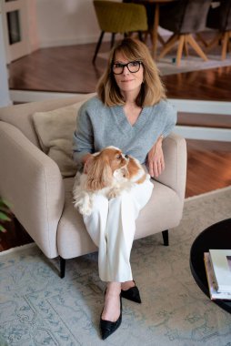 Çekici, orta yaşlı bir kadın sevimli köpeğiyle evdeki koltukta rahatlıyor. Süveter ve pantolon giyen sarışın kadın. Tam boy çekim.  