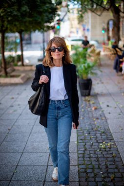Sonbahar günü şehir caddesinde yürüyen olgun, güzel bir kadın. Siyah ceket ve kot giyen sarışın kadın. Tam boy çekim.  