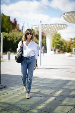 Şehir parkında yürüyen orta yaşlı bir kadın. Beyaz gömlek ve kot giyen sarışın kadın. Tam boy çekim. 